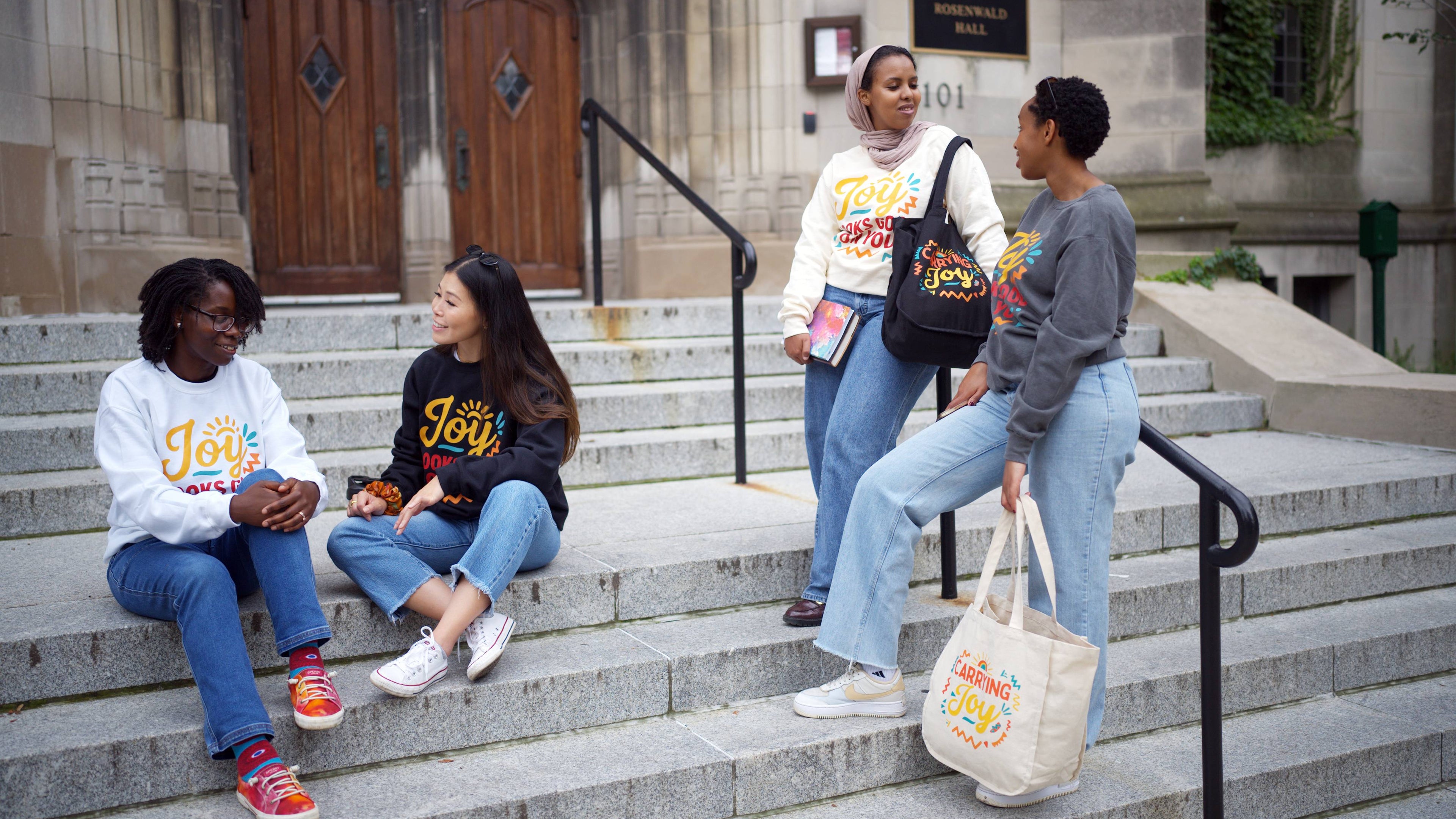 Four women sitting on steps in front of a building, wearing colorful clothing with visible text.